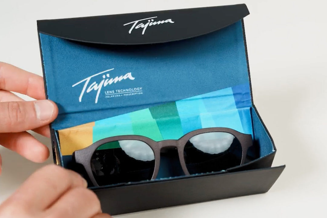 Sunglasses being placed inside an open Tajima foldable case showing blue soft-lined interior and branded microfiber pouch
