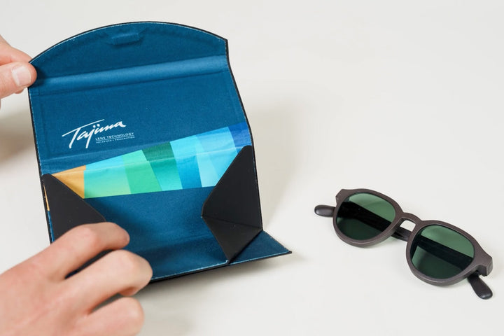 Tajima foldable sunglass case open showing blue branded interior with microfiber pouch next to sunglasses with green polarized lenses